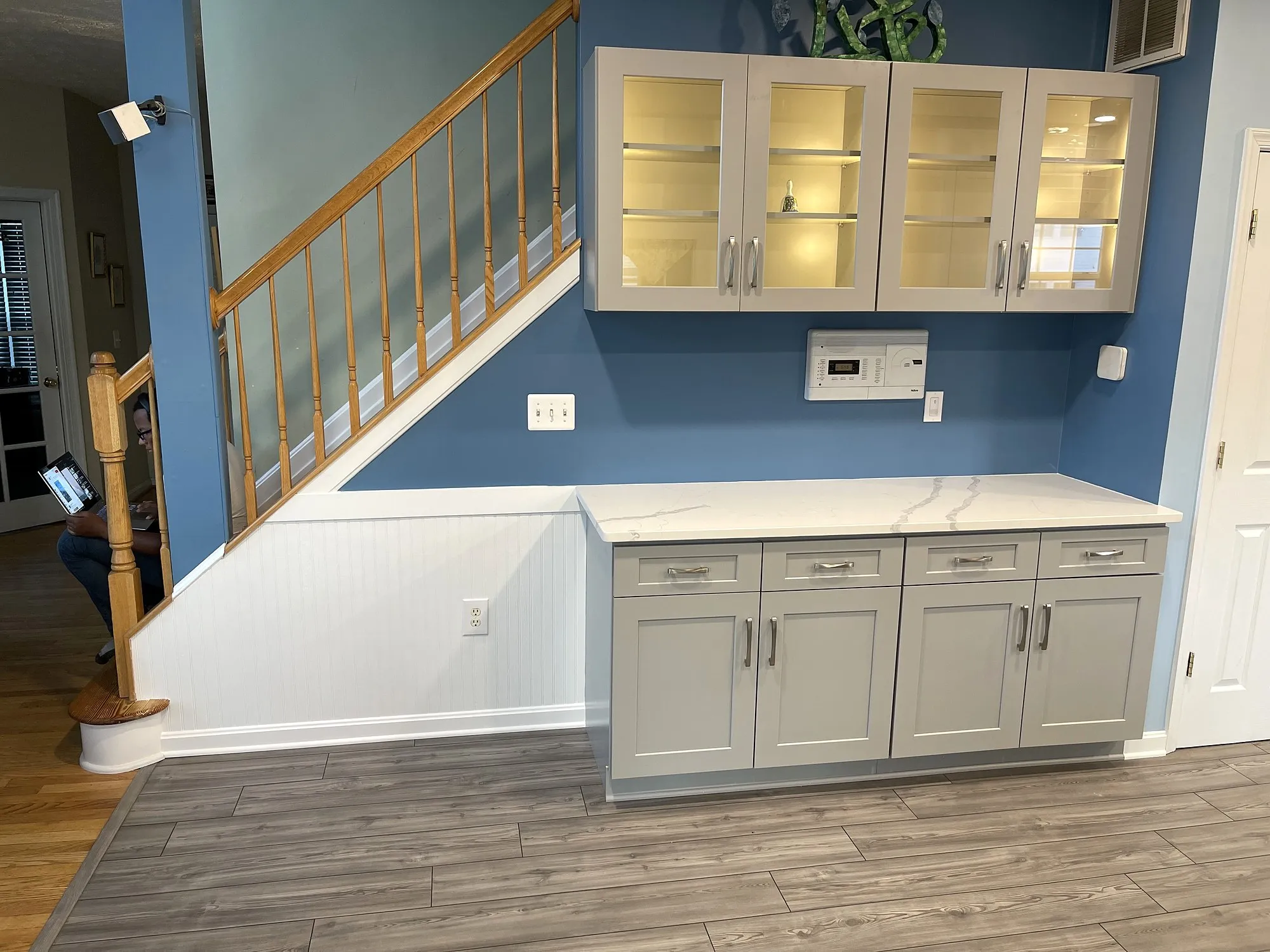 Glass-front upper cabinets and grey base cabinets with a quartz top beside a staircase, beadboard below